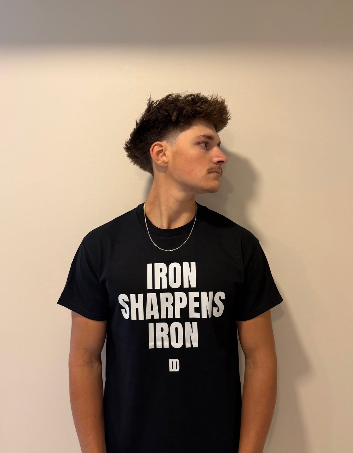 Young man in profile wearing black t-shirt with white text reading Iron Sharpens Iron against beige wall