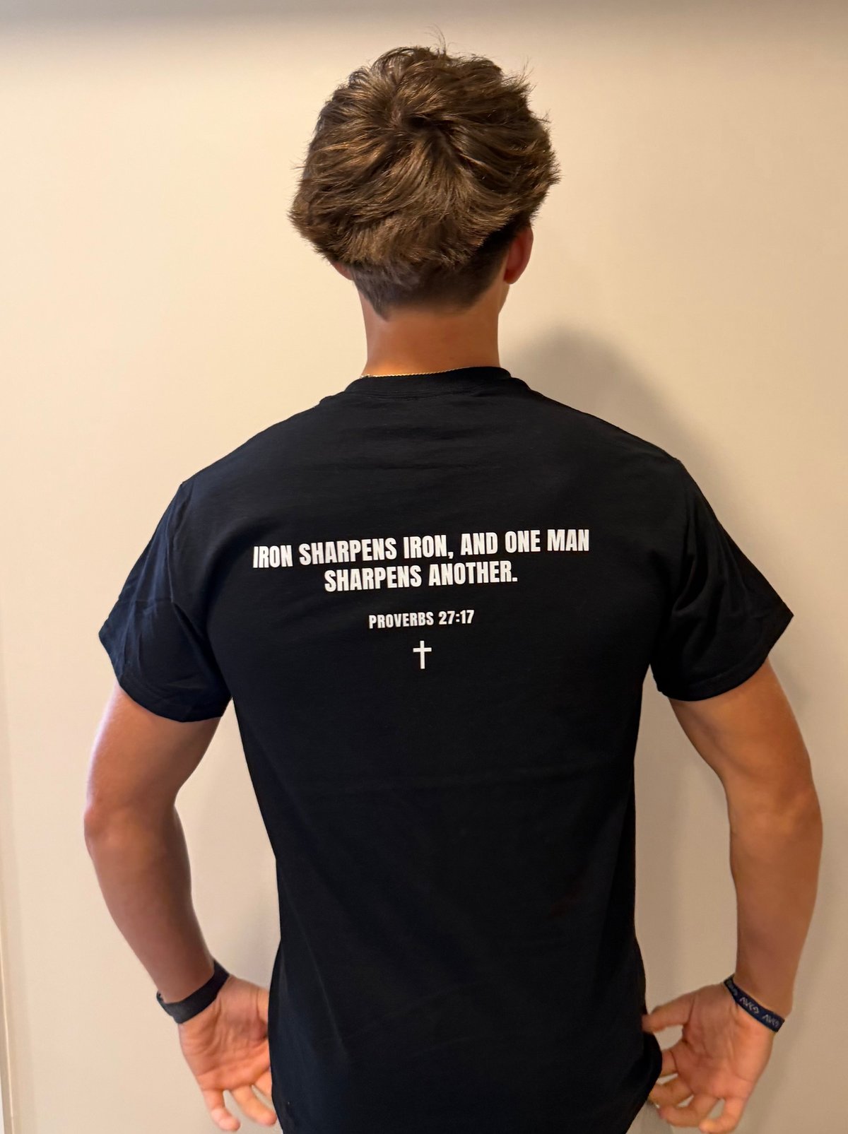 Person wearing a black t-shirt with Proverbs 27:17 printed on the back, standing against a neutral wall