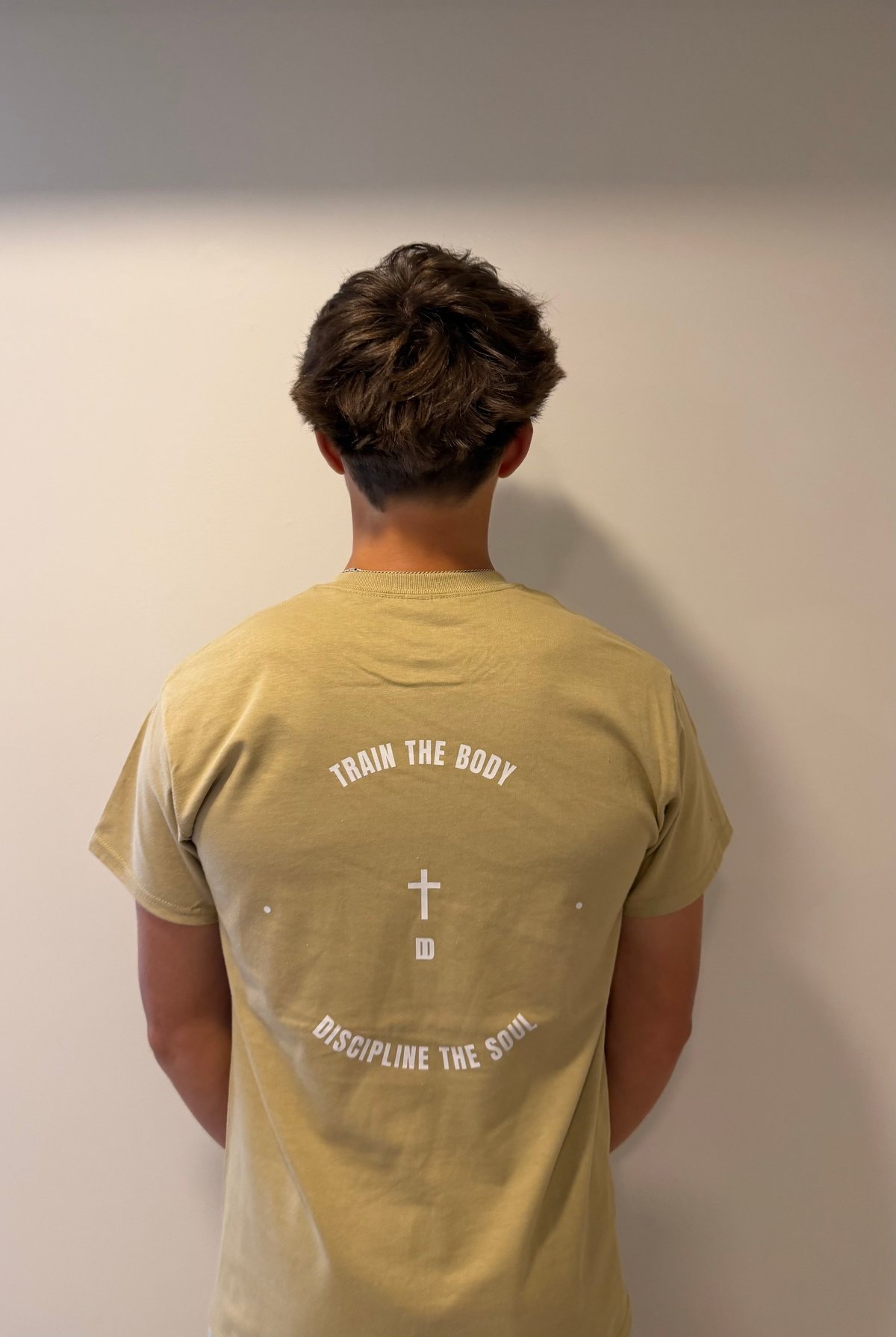 Back view of person wearing mustard yellow t-shirt with text Train the Body and Discipline the Spirit with cross symbol