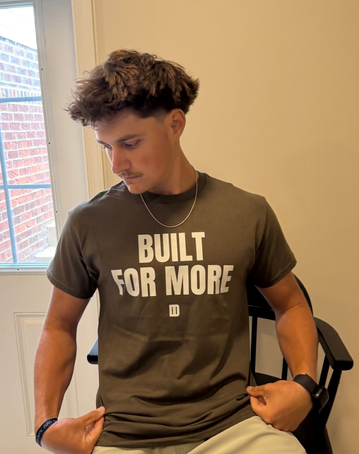Young man wearing olive green t-shirt with Built For More text, seated by window with brick building visible outside