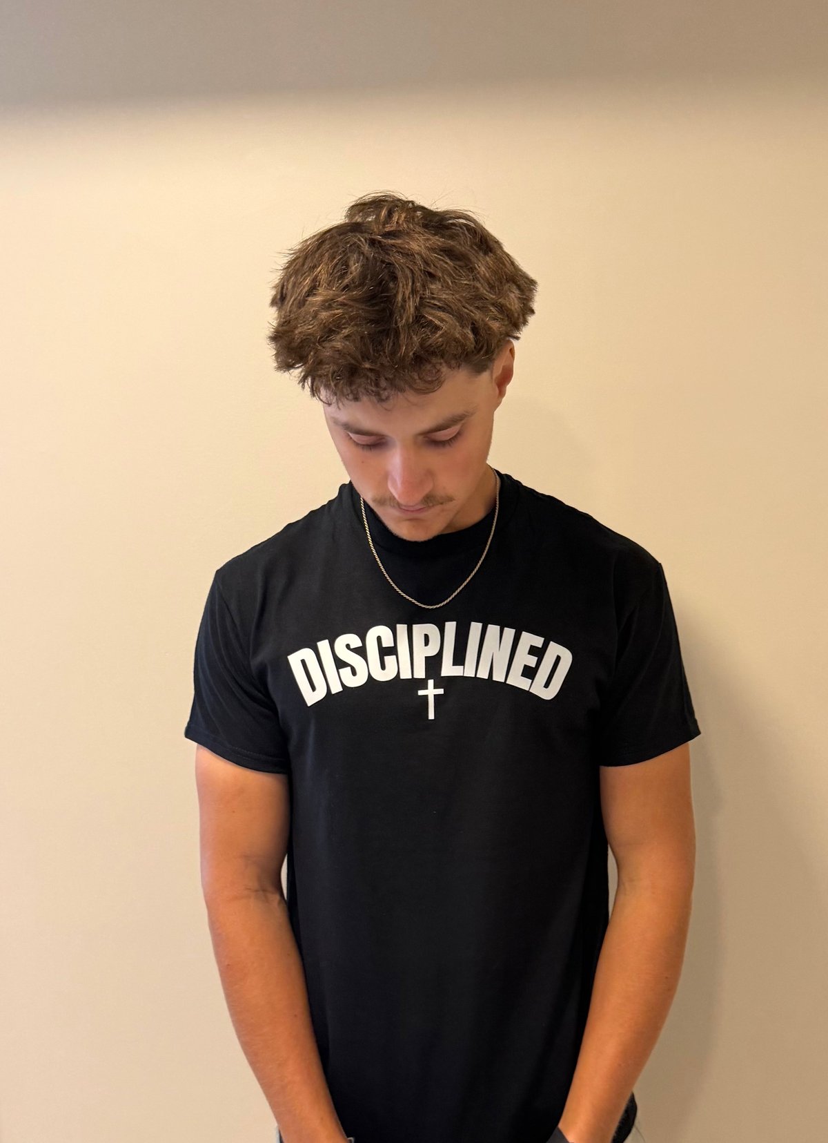 Young man wearing black t-shirt with DISCIPLINED text and cross symbol, looking down against beige background