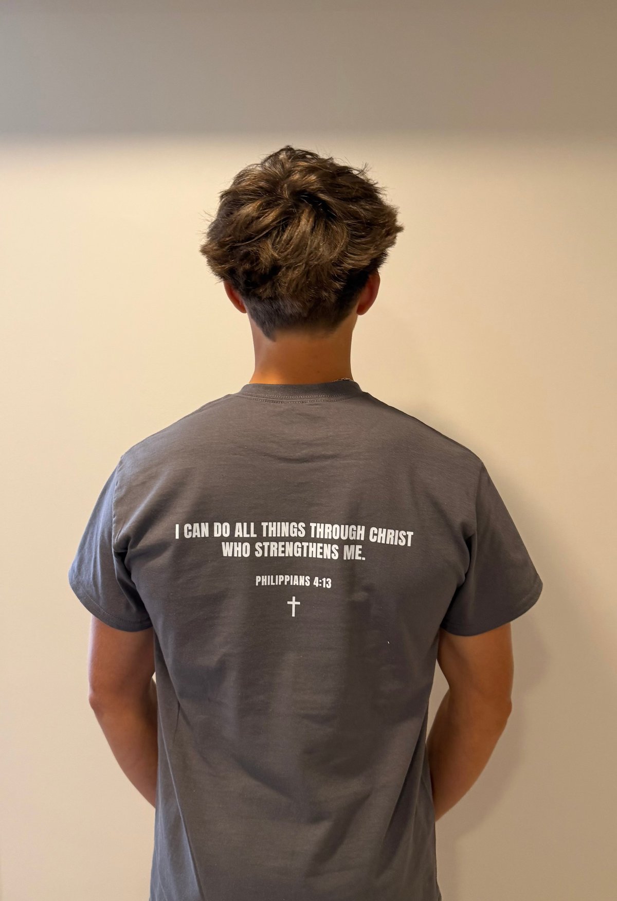 Person wearing gray t-shirt with Philippians 4:13 Bible verse printed on back, standing against neutral wall