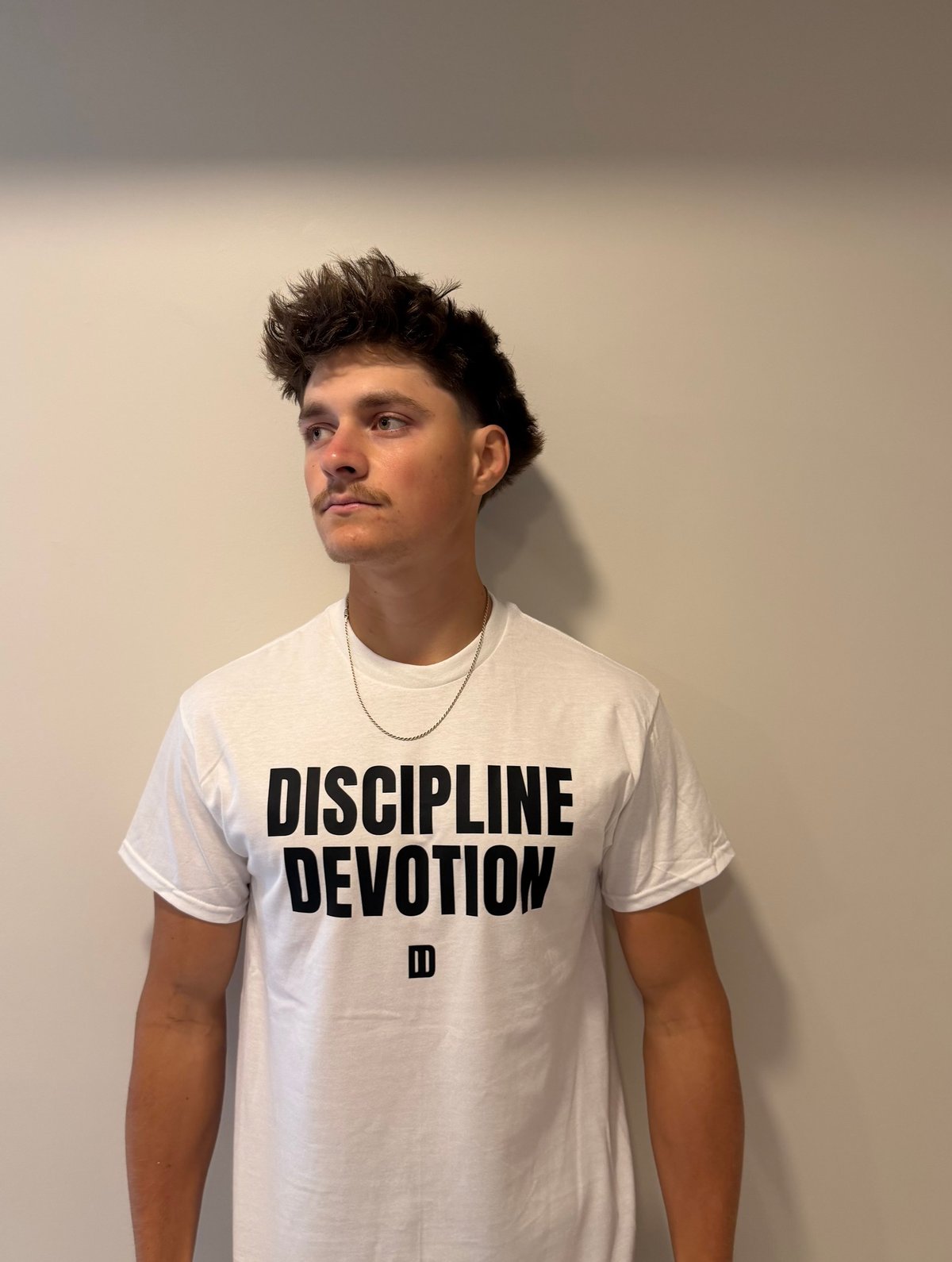 Man wearing white Discipline Devotion t-shirt with chain necklace against beige wall