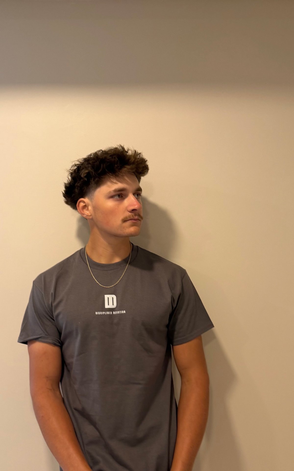 Young man wearing a dark gray t-shirt and silver chain necklace, posed against a beige wall looking to the side
