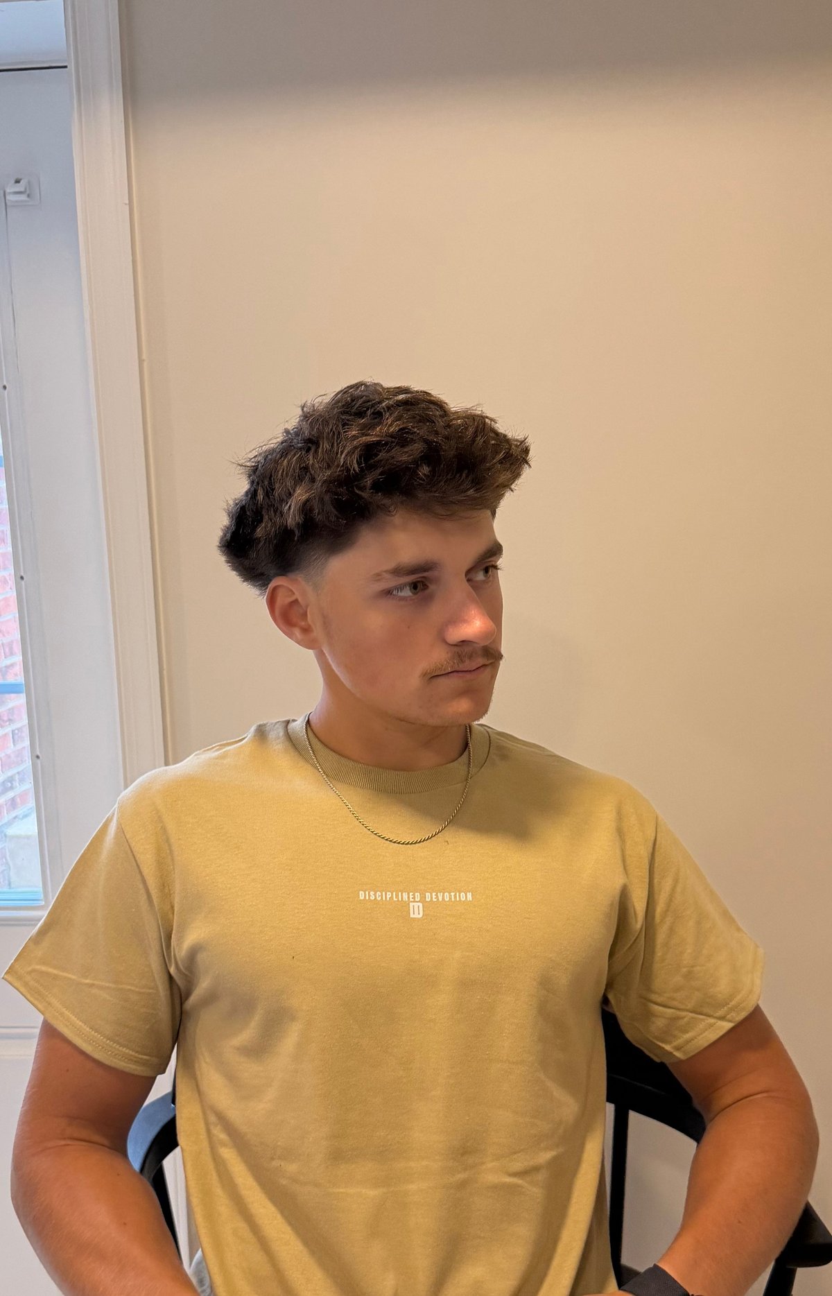 Young man with textured dark hair wearing a mustard yellow graphic t-shirt and chain necklace, indoors against a neutral beige wall