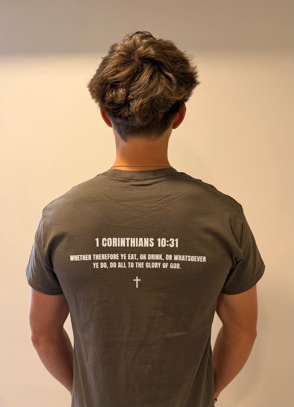 Person wearing gray t-shirt with 1 Corinthians 10:31 Bible verse and cross printed on back, standing against neutral background
