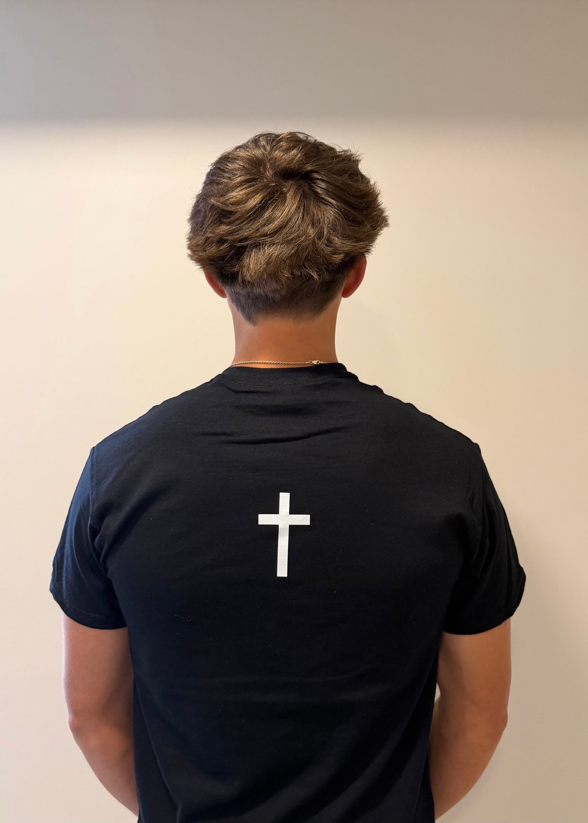 Person wearing black t-shirt with white cross symbol on back, viewed from behind against neutral beige background
