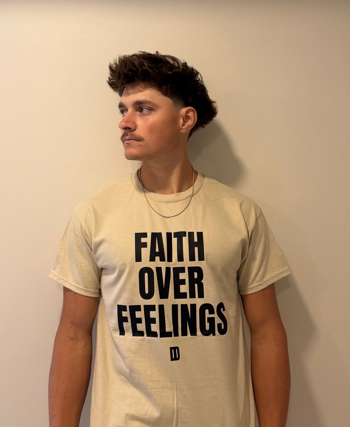 Young man with dark hair wearing a cream-colored t-shirt that reads FAITH OVER FEELINGS, standing against a beige wall