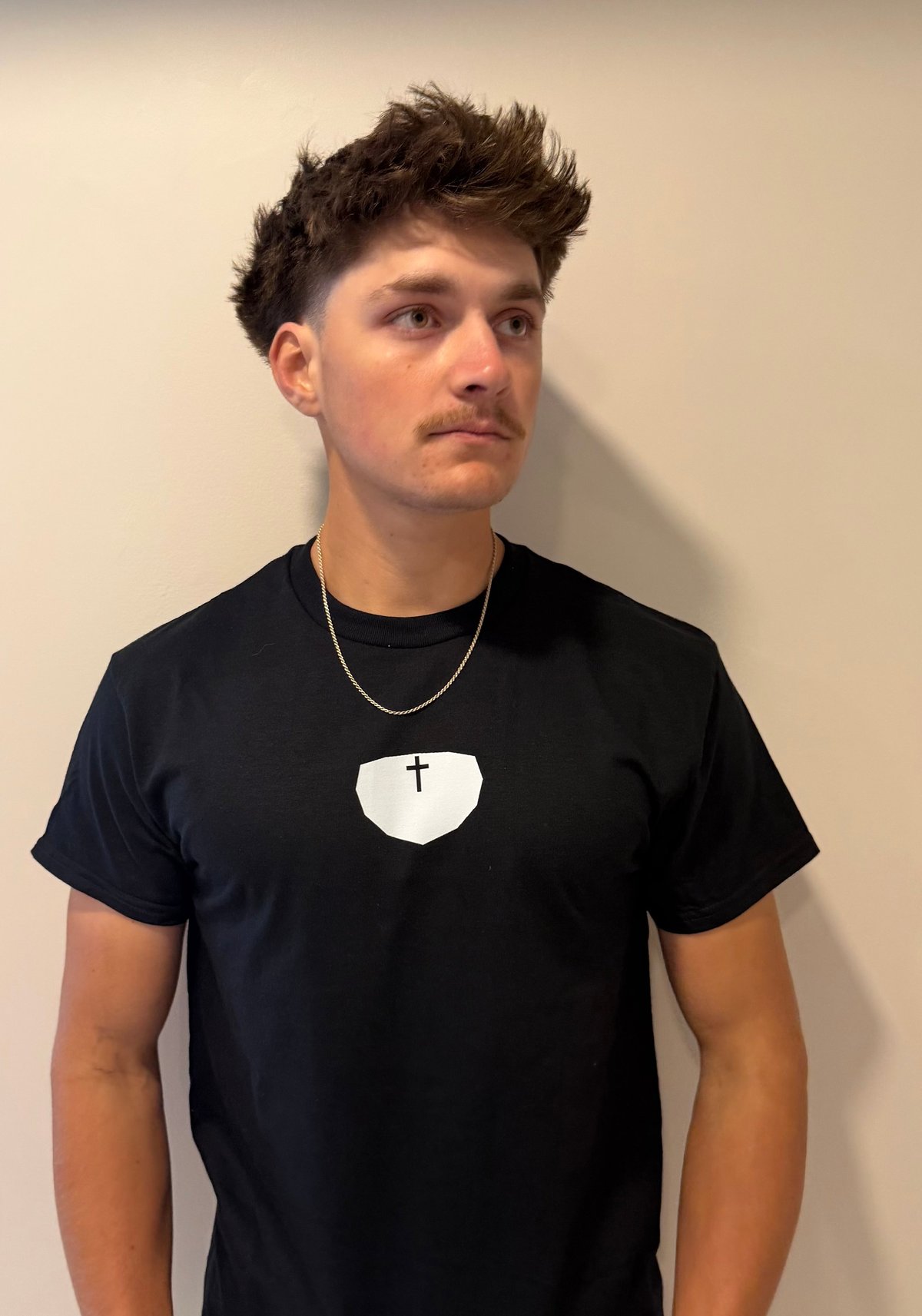 Young man with dark styled hair wearing a black t-shirt with a white cross design and silver chain necklace against a neutral background