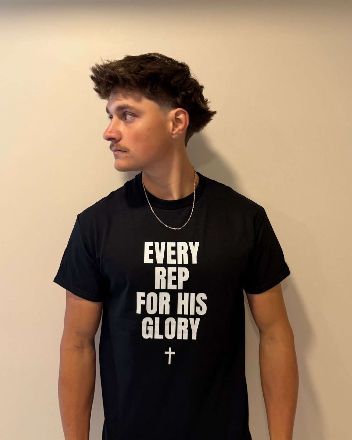 Young man wearing black t-shirt with white text reading EVERY REP FOR HIS GLORY and a cross symbol, standing against a beige wall
