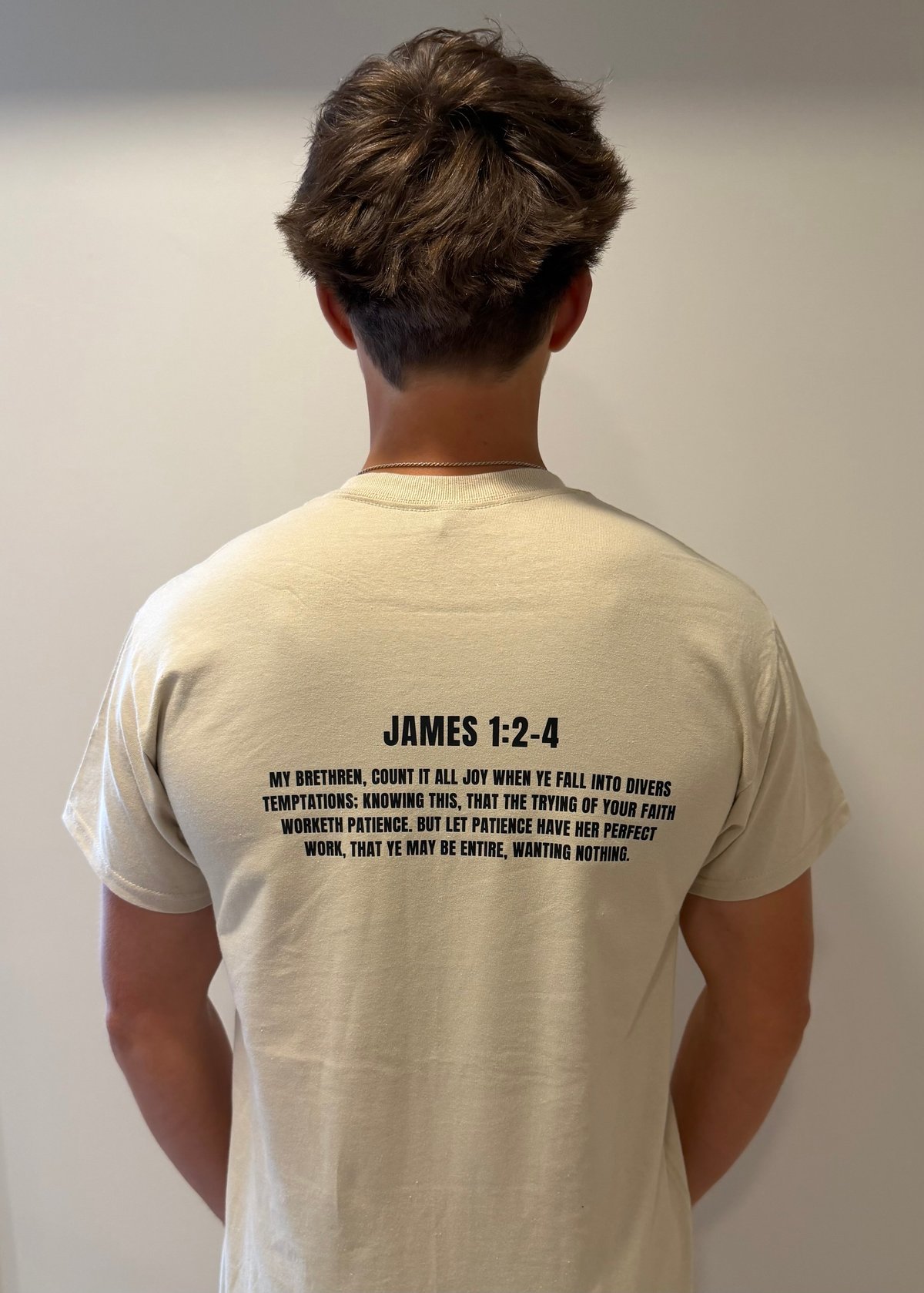 Person wearing cream-colored t-shirt with James 1:2-4 Bible verse printed on back, photographed from behind against neutral background