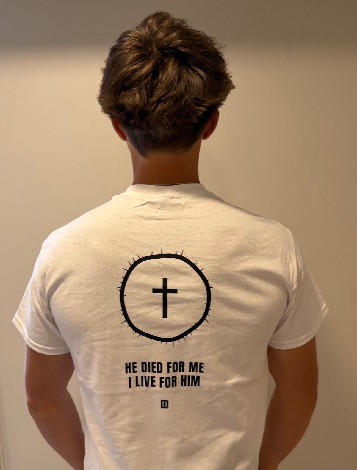 Person wearing white t-shirt with circular cross design and text He Died For Me I Live For Him printed on back