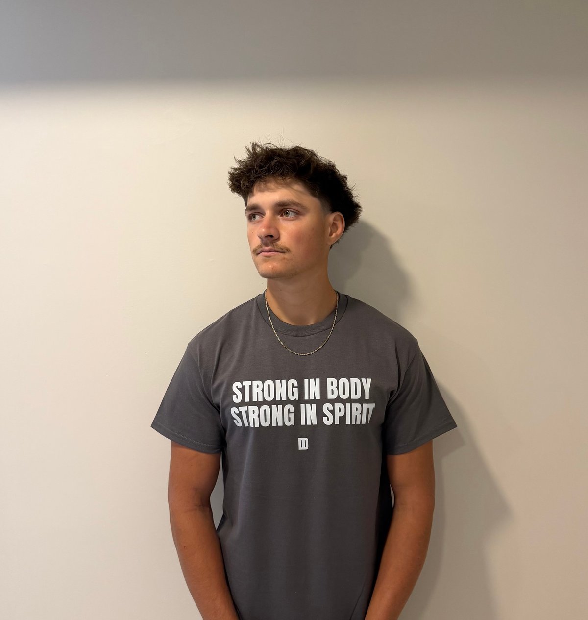 Young man wearing a dark gray t-shirt with white text reading Strong in Body Strong in Spirit standing against a plain white wall