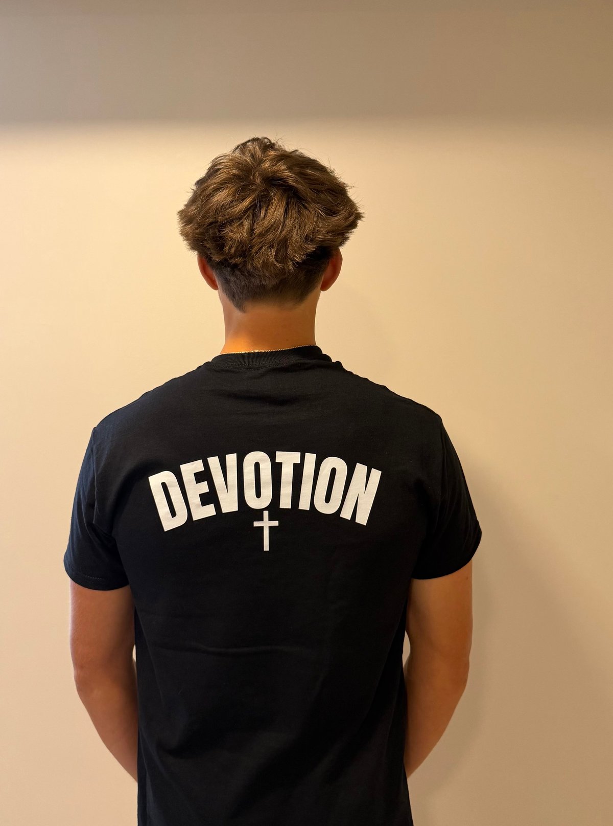 Person wearing black t-shirt with white DEVOTION text and cross symbol, viewed from behind against beige wall