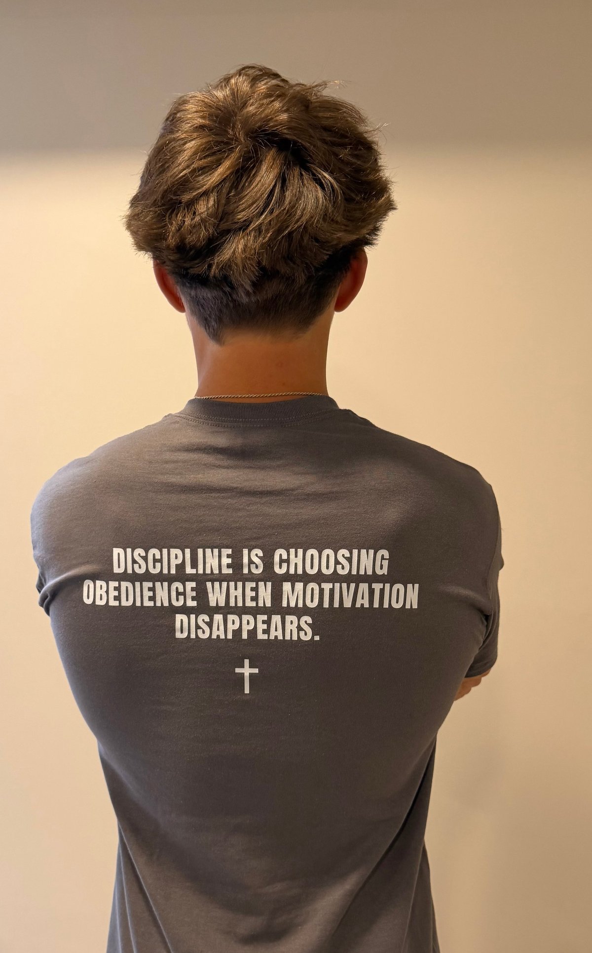 Person viewed from behind wearing a gray t-shirt with text reading Discipline is choosing obedience when motivation disappears and a cross symbol